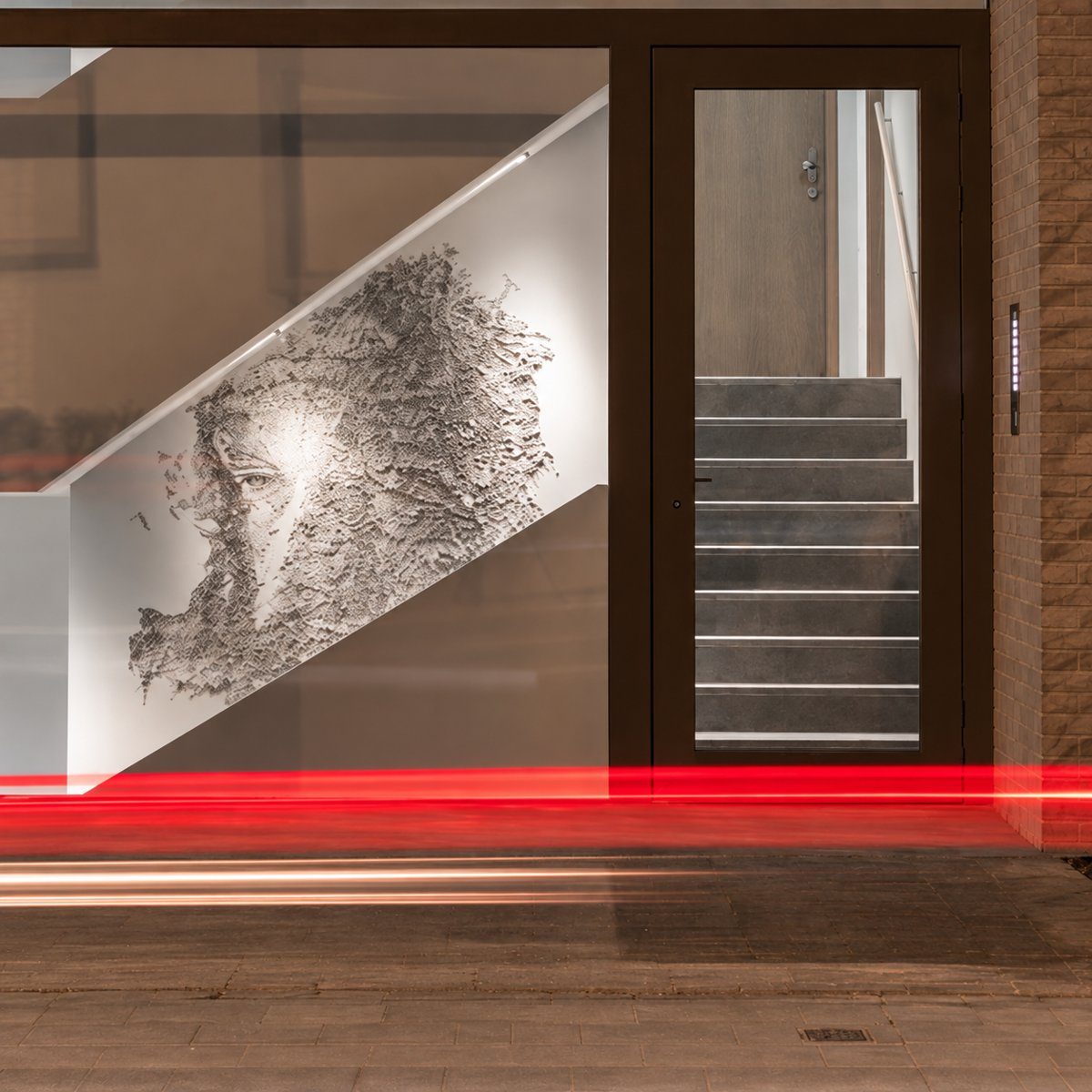 A face carved into a white stairwell wall by Vhils, seen through the glass entrance at night