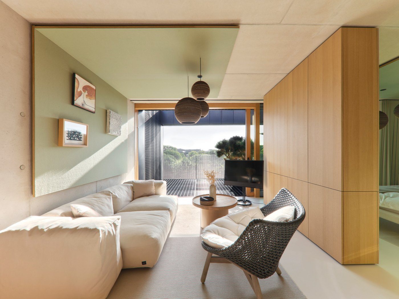 Rise, a short-stay apartment in warm sunlight: a linen sofa under a green ceiling panel, a woven armchair, a tall oak wall and a terrace beyond