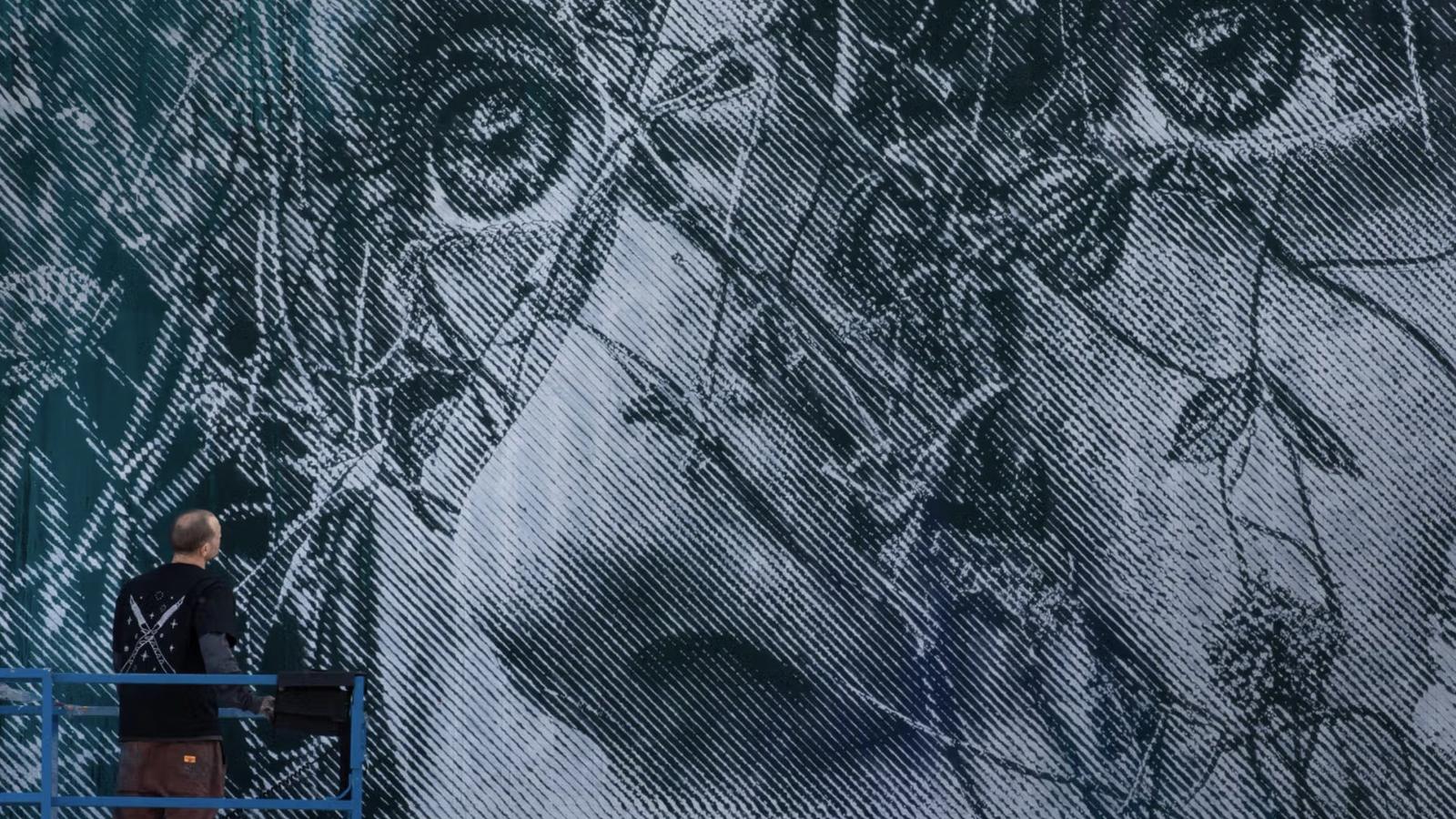 A giant stencil mural by SNIK at LARGO: two faces behind leaves in fine diagonal lines, the artist on a lift in front of it
