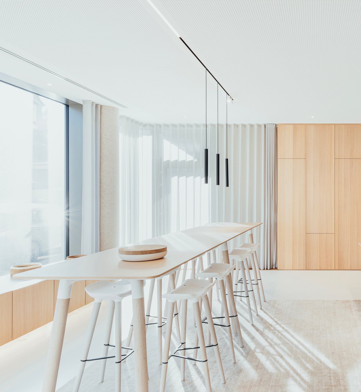 A bright interior with a long oak table, white stools, an acoustic ceiling and floor-length curtains