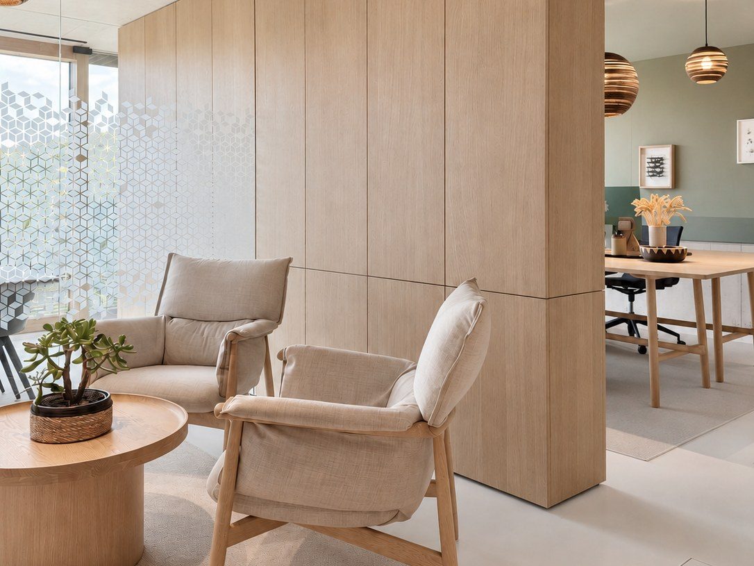 The Stik office lounge: two linen armchairs and a round oak table in front of a tall oak wall, patterned glass and a view into a green-walled workroom